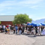 A procession of families and students winds through Farm and Ranch Museum grounds with barn and mountains in the background