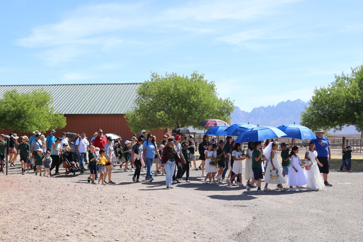 A procession of families and students winds through Farm and Ranch Museum grounds with barn and mountains in the background
